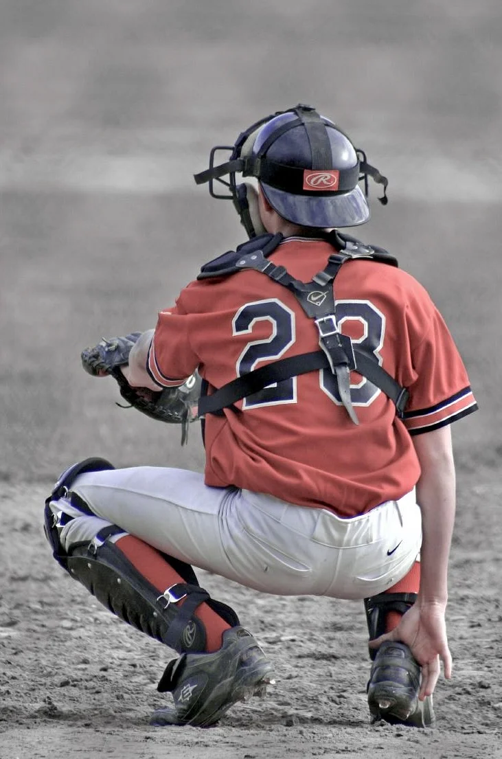A baseball catcher in a red jersey numbered 28, seen from behind in a crouch with mask, chest protector, and shin guards on a dirt field.