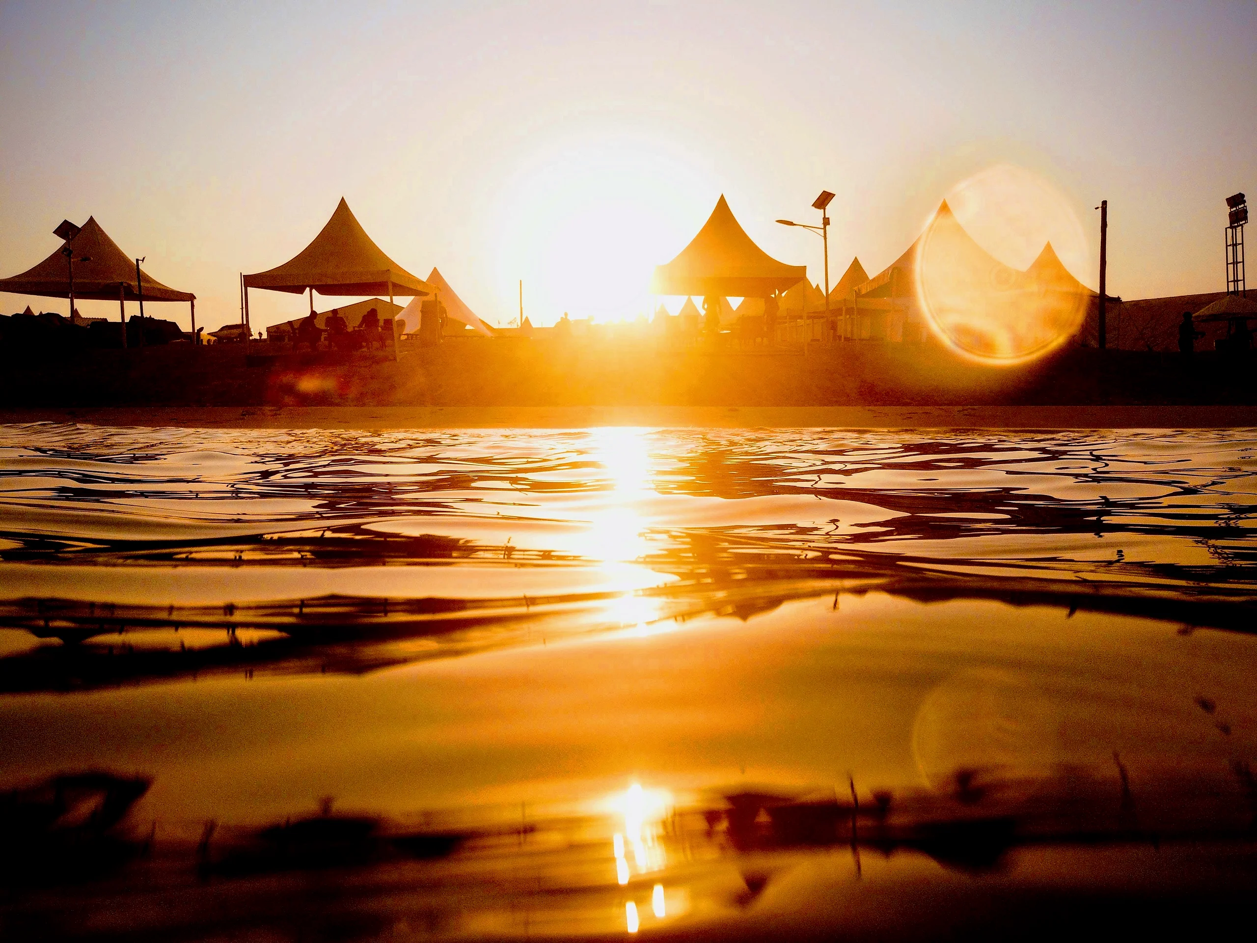 Row of peaked tents silhouetted against a low sun on a shoreline, with the light mirrored across rippled water in the foreground.
