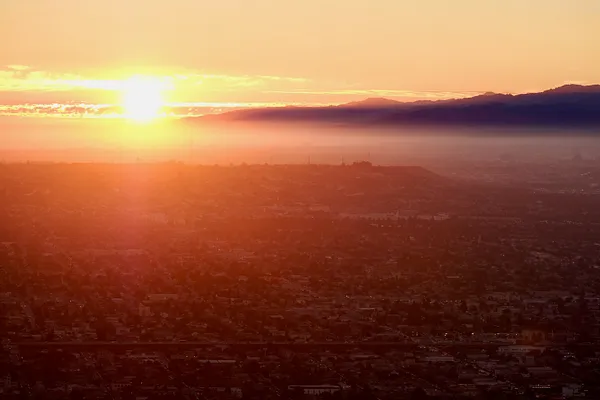 The sun rises over a hazy Los Angeles basin, its low sprawl fading into distant mountains.
