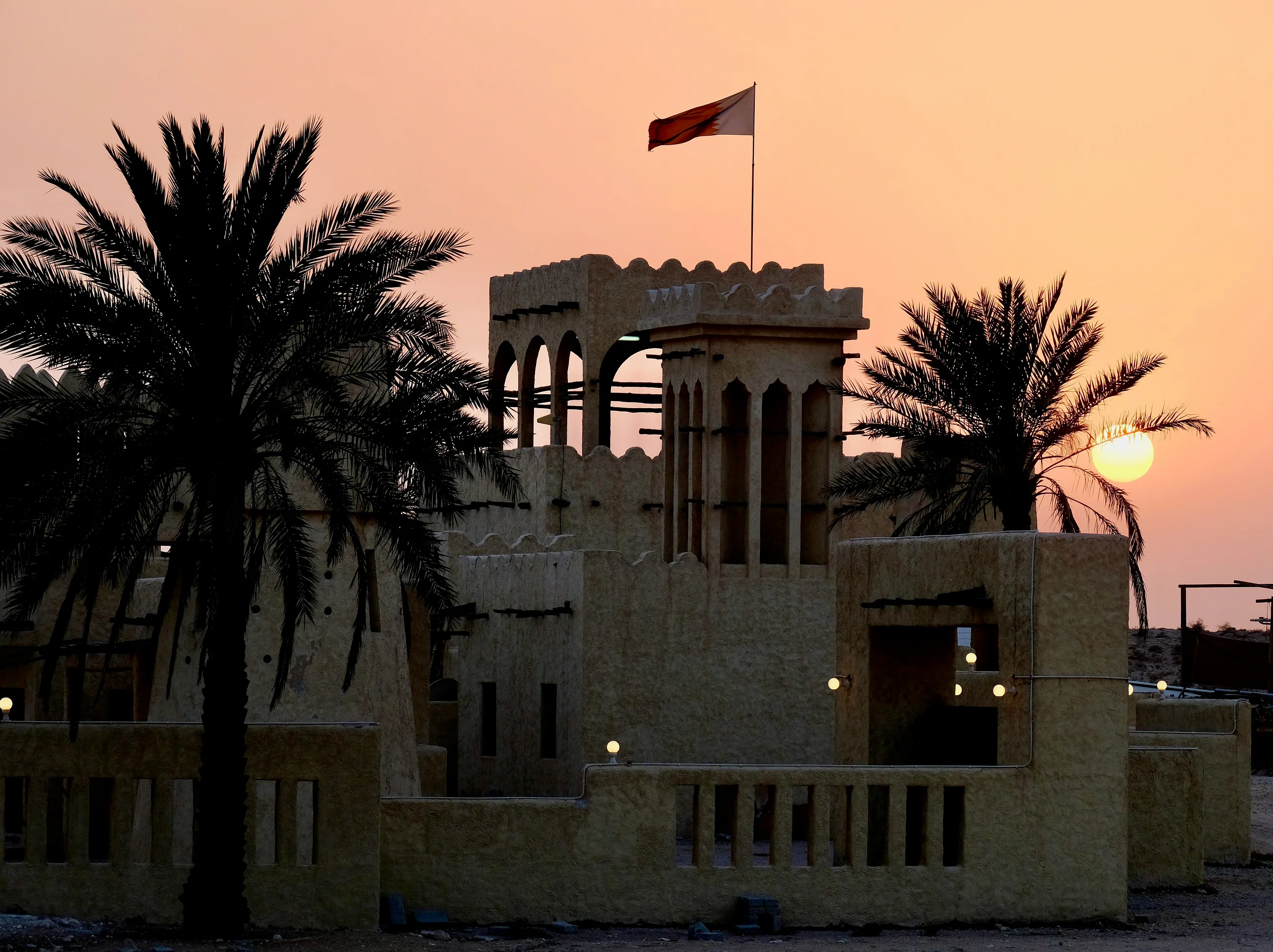 A sand-colored windtower flying the Qatari flag stands behind silhouetted date palms as the sun sets in a peach sky.