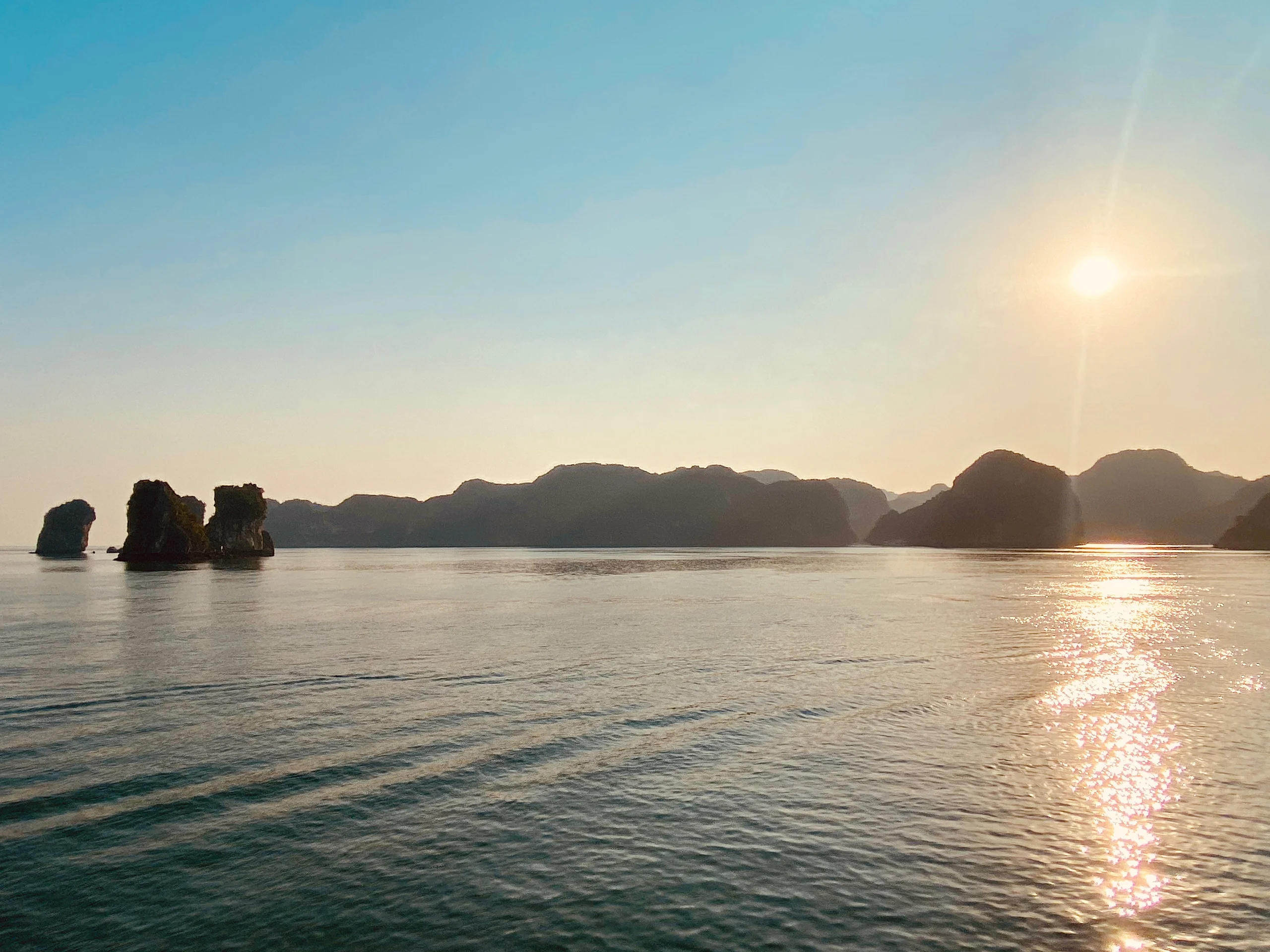 Limestone karst islands sit low on the horizon of a calm bay while the sun burns bright at right, laying a path of light across the water.