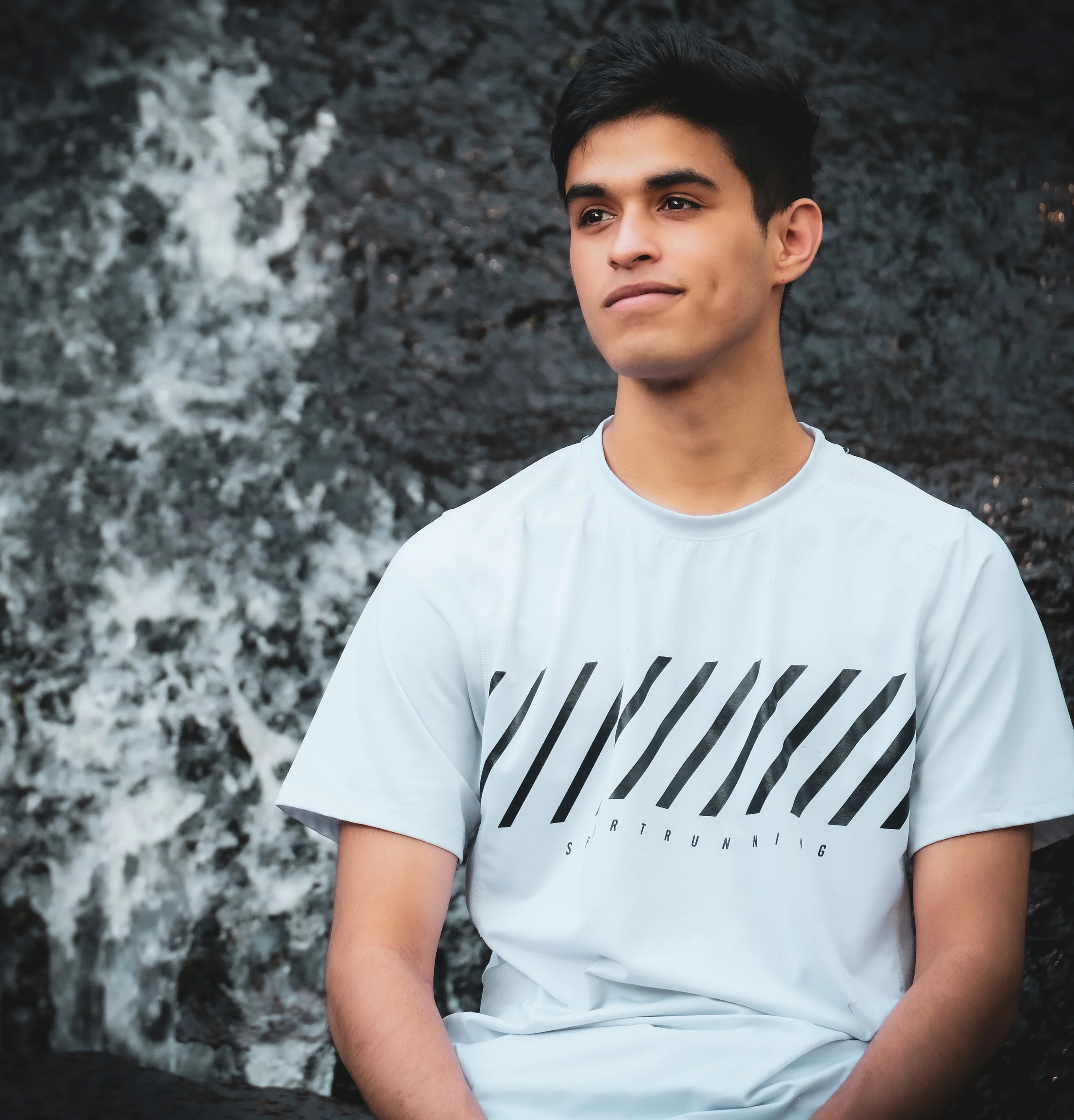 A young man in a white running shirt sits before a dark, water-streaked rock face, looking off to the side.