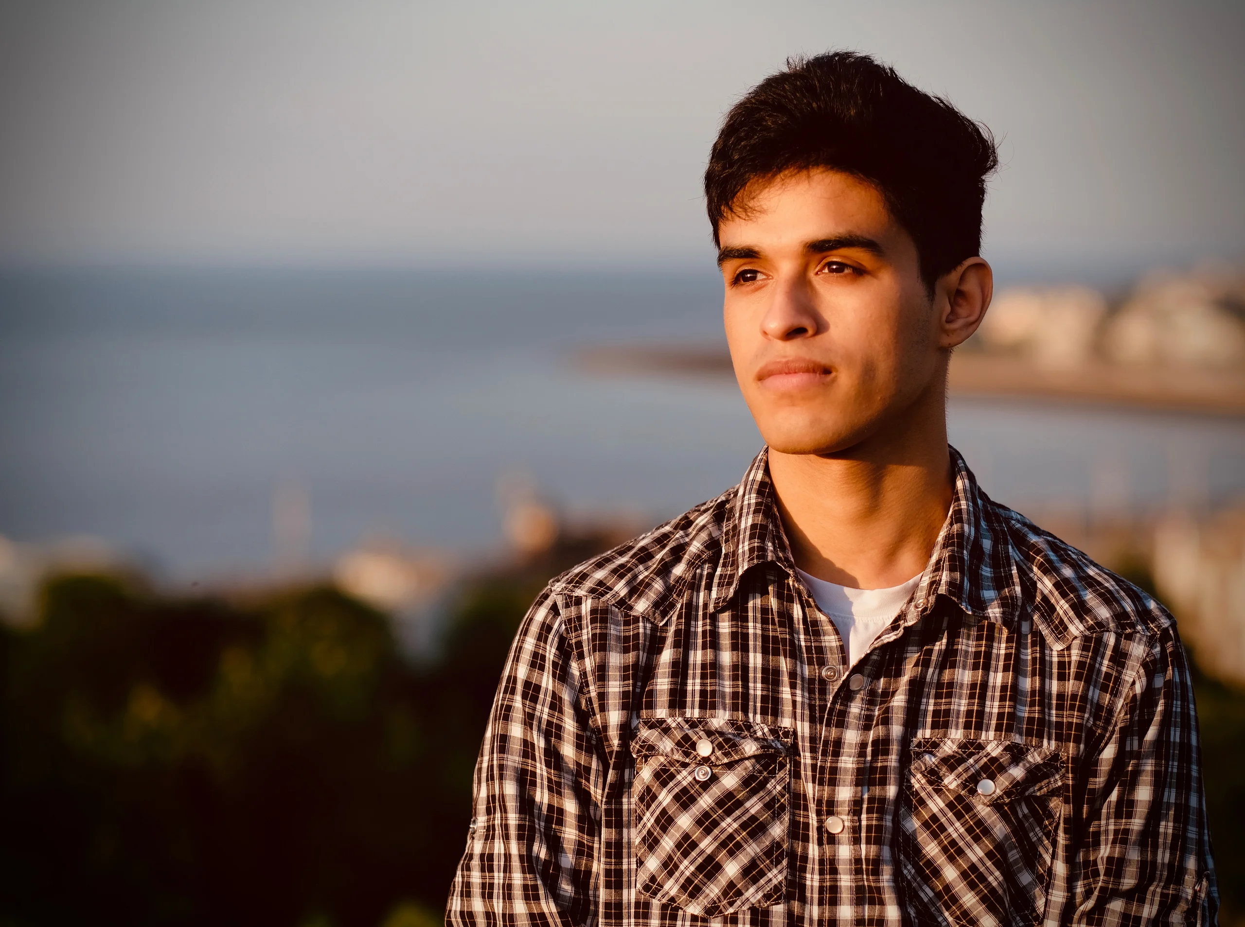 A young man in a plaid shirt looks off to the side, lit by warm low sun, with a blurred sea and distant shoreline behind him.