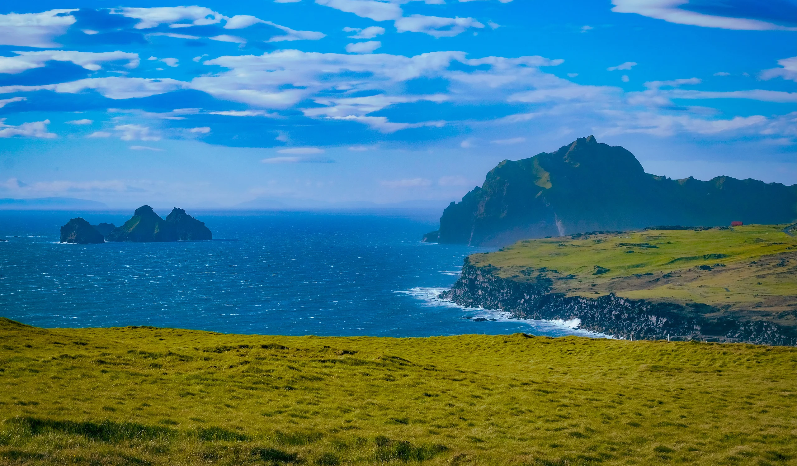 Grassy headland sloping to sea cliffs with a jagged green peak and small rocky islets under a bright blue sky.