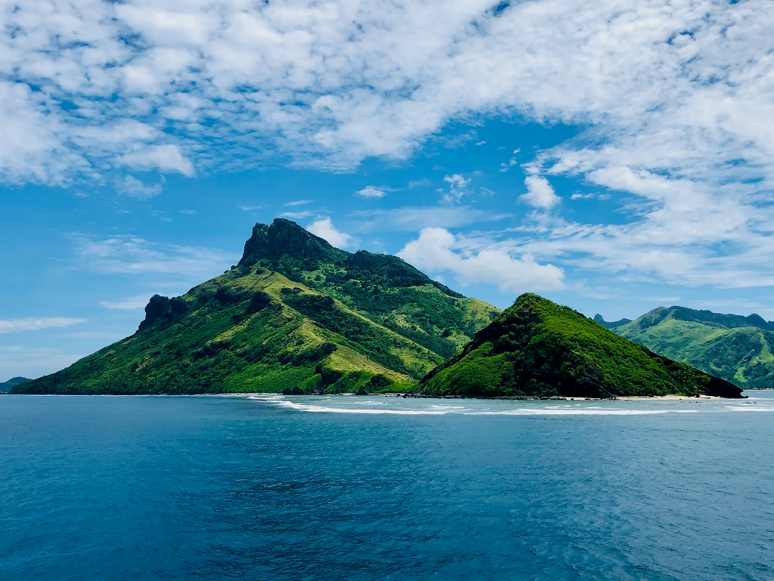 A green, twin-peaked island rises from a turquoise sea beneath a broad blue sky scattered with clouds.
