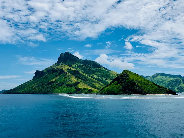 A green, twin-peaked island rises from a turquoise sea beneath a broad blue sky scattered with clouds.