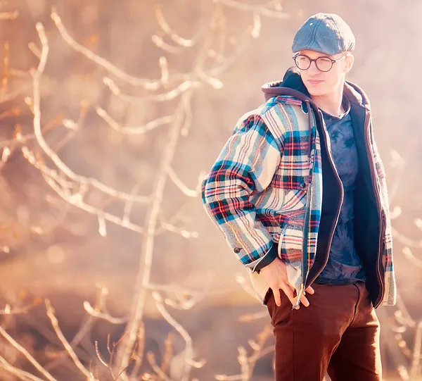 A man in a flat cap and round glasses wears a bright plaid wool coat, hand on hip, before a hazy backlit thicket of bare branches.