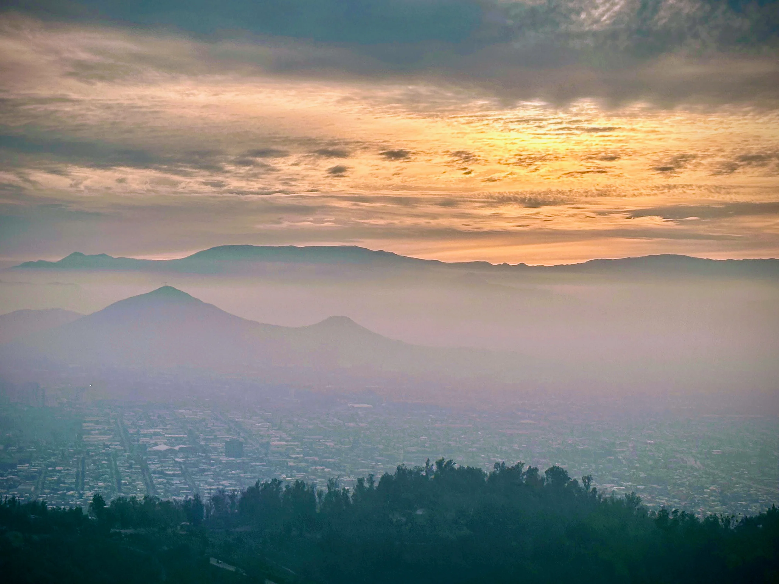A hazy city sprawls across a valley beneath layered mountain ridges under a cloudy sunset sky.