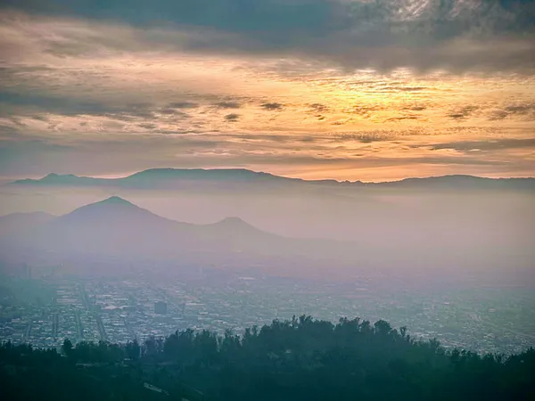 A hazy city sprawls across a valley beneath layered mountain ridges under a cloudy sunset sky.