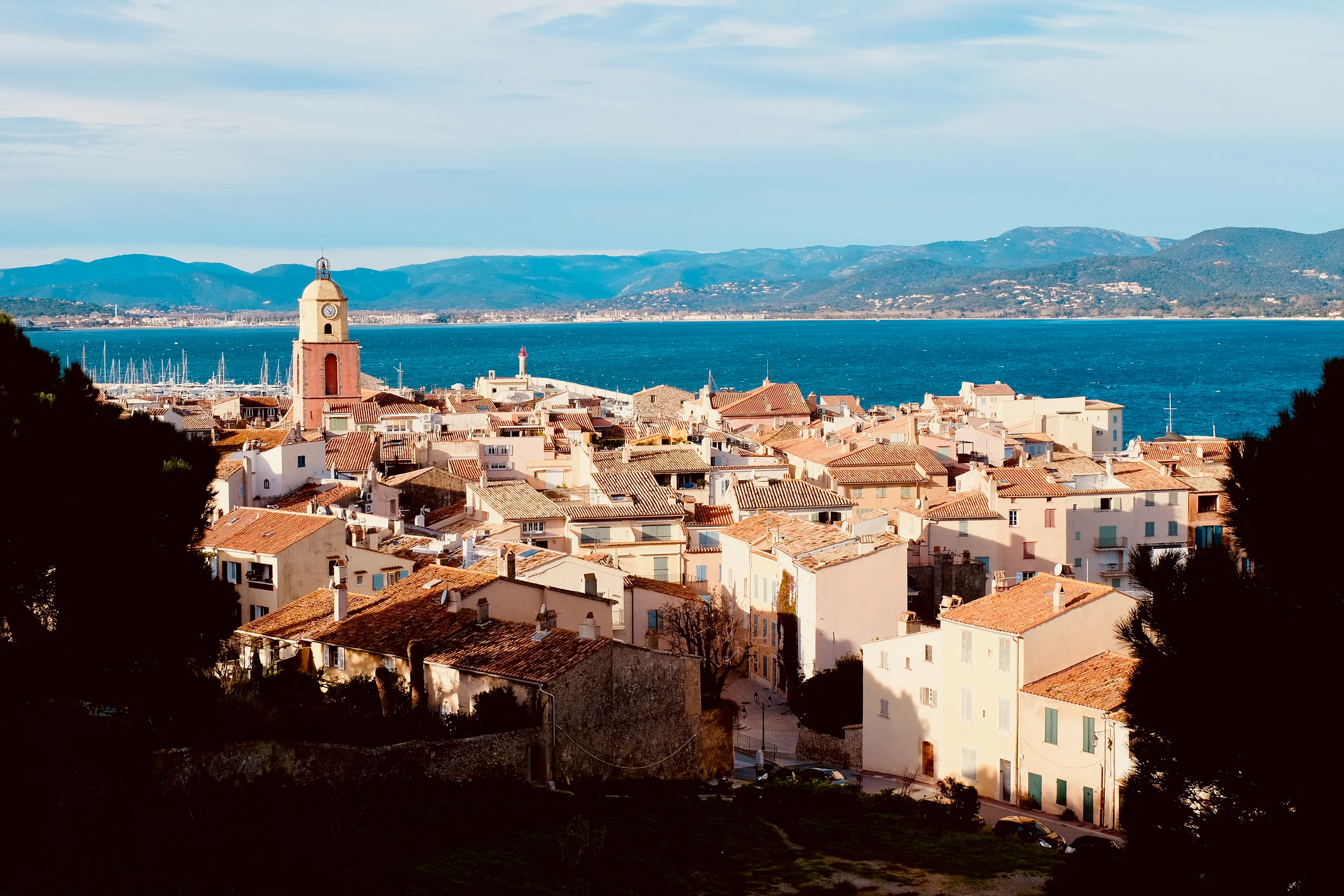 Terracotta rooftops of Saint-Tropez cluster around a bell tower above a deep blue bay backed by distant hills.