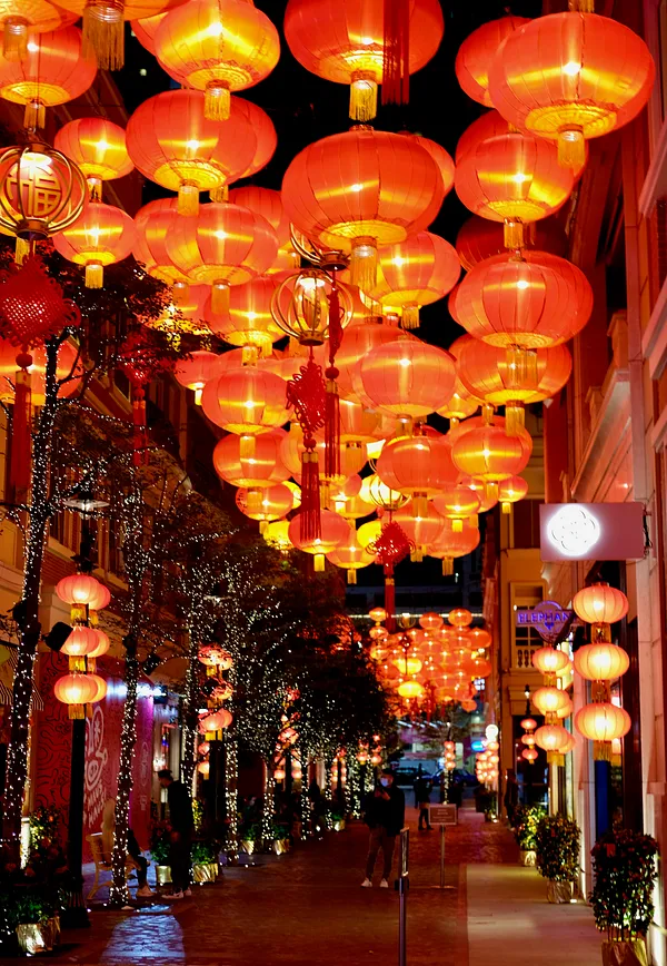 A pedestrian walkway at night crowded overhead with dozens of glowing red-and-gold lanterns, with light-wrapped trees and a lone figure walking the length of the lane.