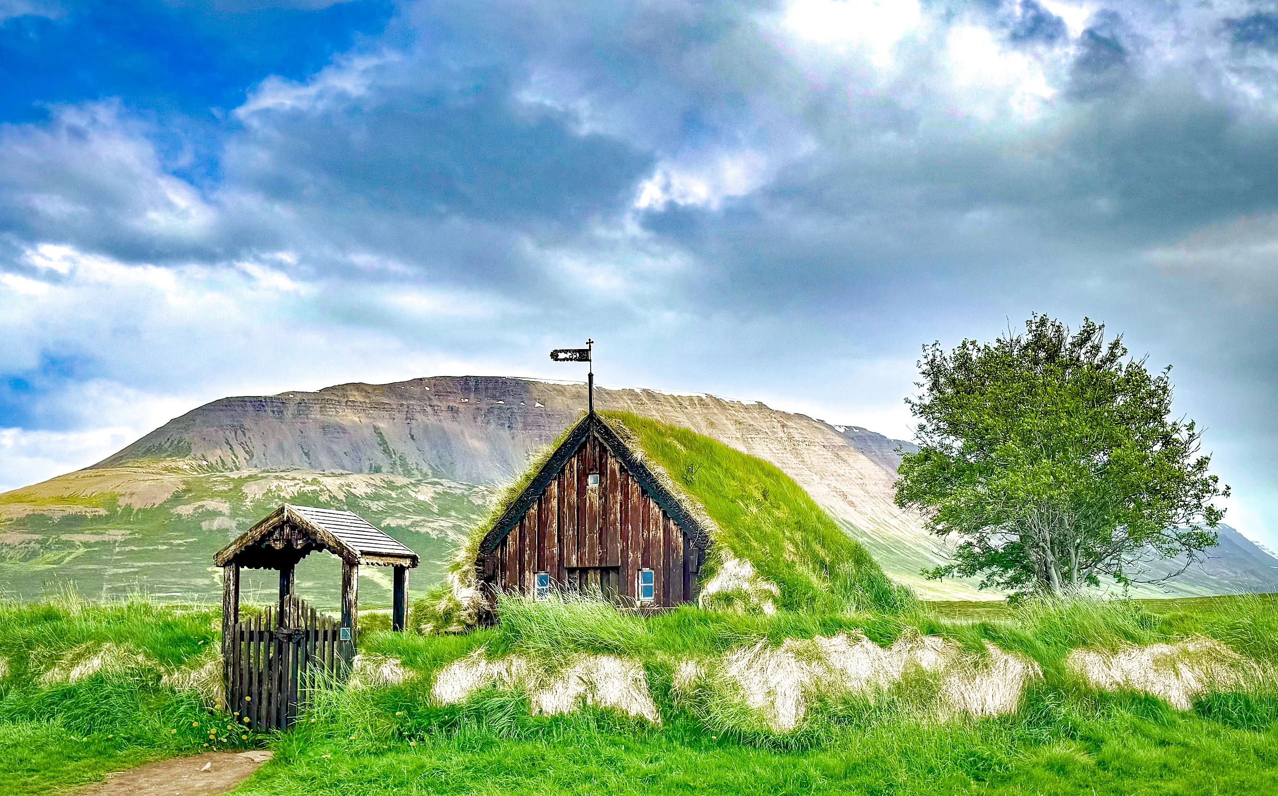 A wooden turf-roofed church with a grassed gate stands in a green meadow below a broad mountain and a cloudy sky.