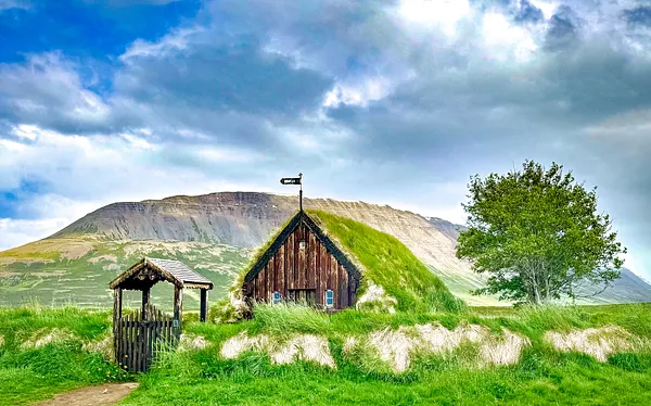 A wooden turf-roofed church with a grassed gate stands in a green meadow below a broad mountain and a cloudy sky.