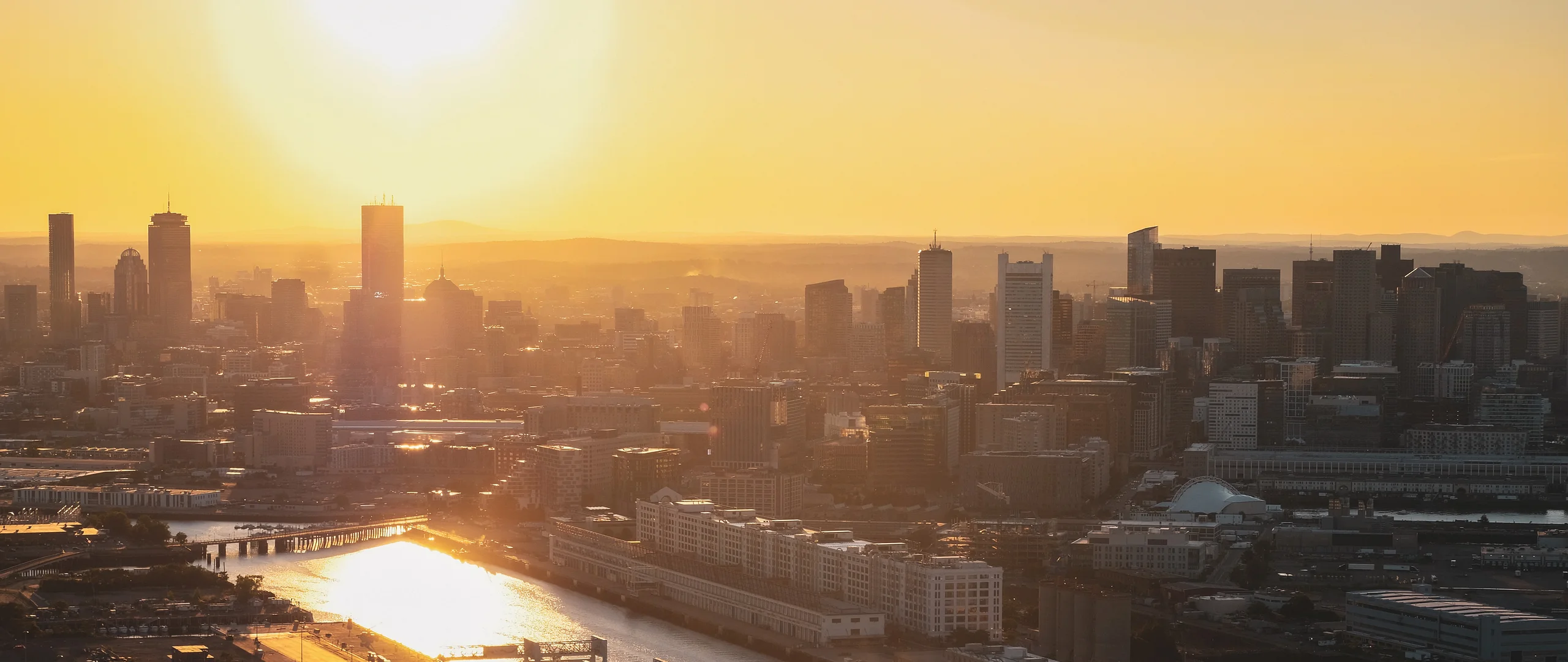 A city skyline seen from above at sunrise, with the sun low over the buildings and its light reflecting off a river in the foreground.
