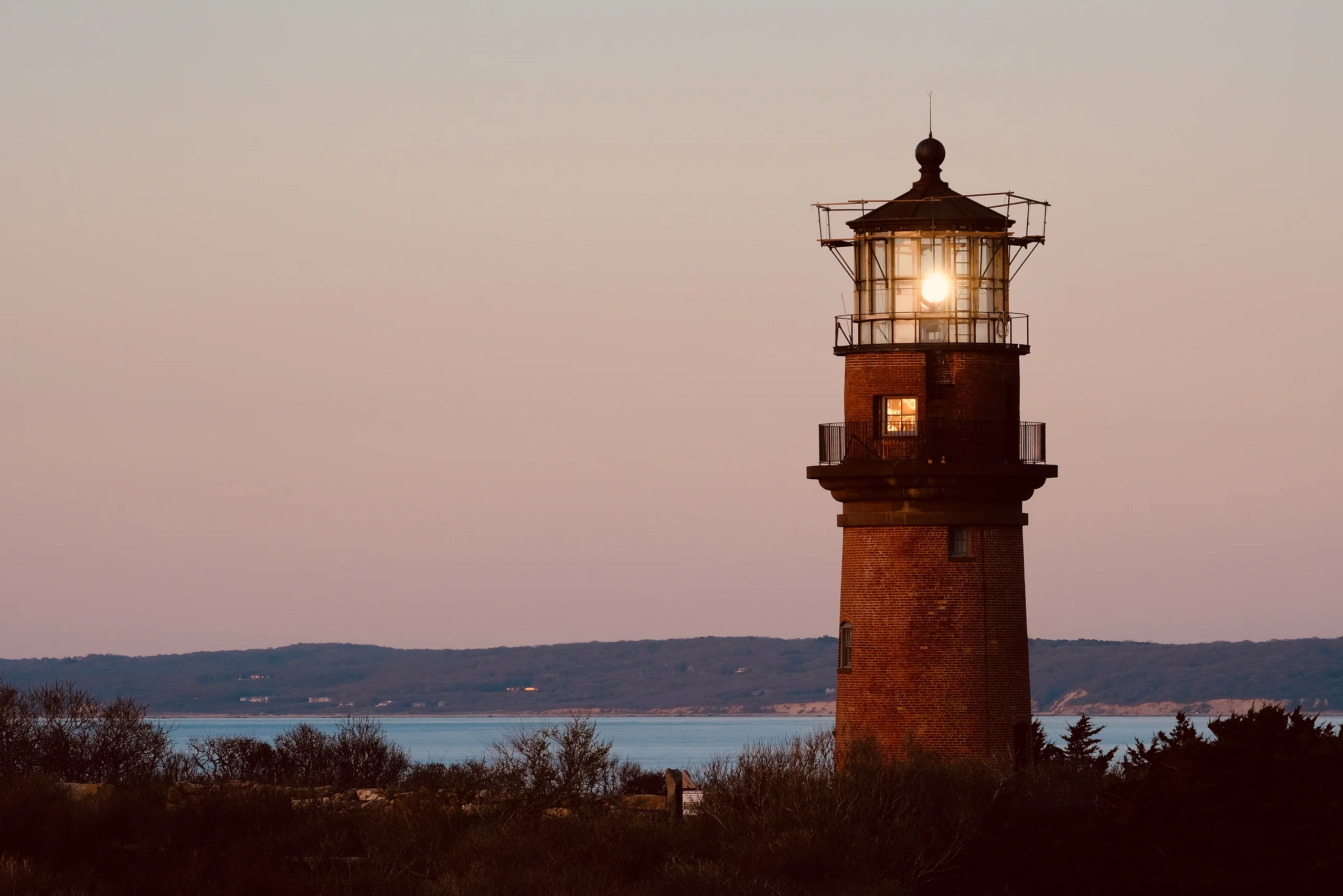 A brick lighthouse with its lamp lit stands above low brush at dusk, the sea and distant hills fading behind it under a rose-colored sky.