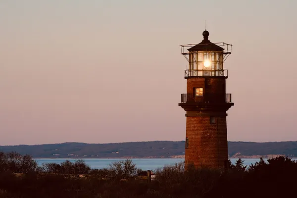 A brick lighthouse with its lamp lit stands above low brush at dusk, the sea and distant hills fading behind it under a rose-colored sky.