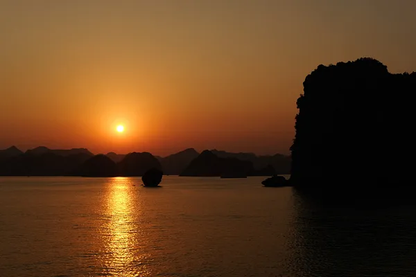 The sun sets over Ha Long Bay, casting a golden path across the water between silhouetted limestone karsts.