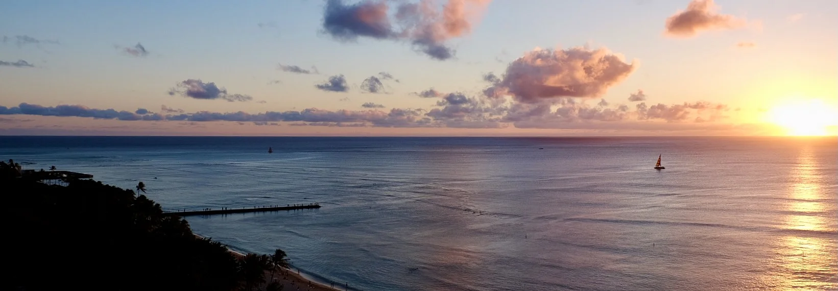 A calm sea at sunset with a small sailboat on the horizon, a long pier reaching from a shadowed shore, and the sun low behind scattered clouds.