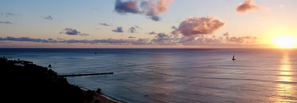A calm sea at sunset with a small sailboat on the horizon, a long pier reaching from a shadowed shore, and the sun low behind scattered clouds.