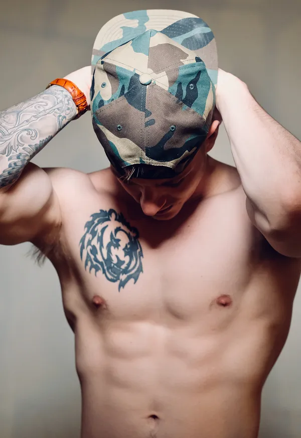 A shirtless man with a tiger tattoo on his chest and a sleeve tattoo on one arm tilts his head down while adjusting a camouflage cap against a plain gray backdrop.