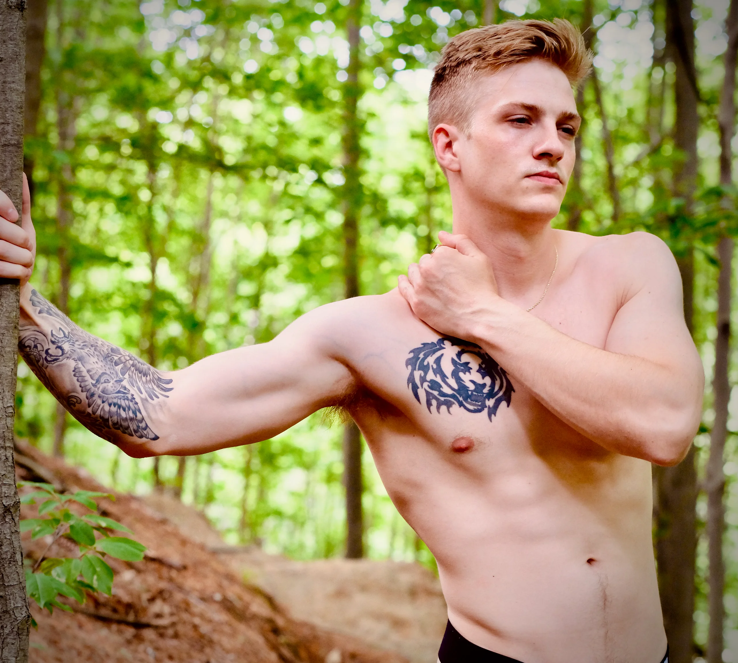 A shirtless young man with tattoos on his arm and chest stands in a sunlit forest, one hand resting on his shoulder, the other braced against a tree.