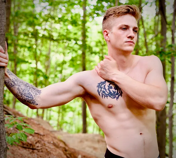 A shirtless young man with tattoos on his arm and chest stands in a sunlit forest, one hand resting on his shoulder, the other braced against a tree.