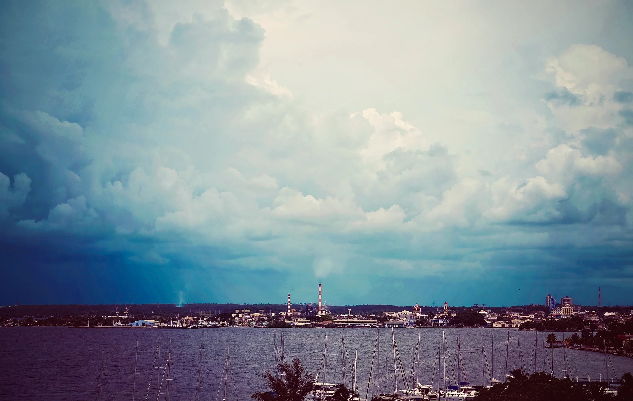 A harbor town seen across the bay under towering clouds, with sailboat masts in the foreground and twin smokestacks on the far shore.