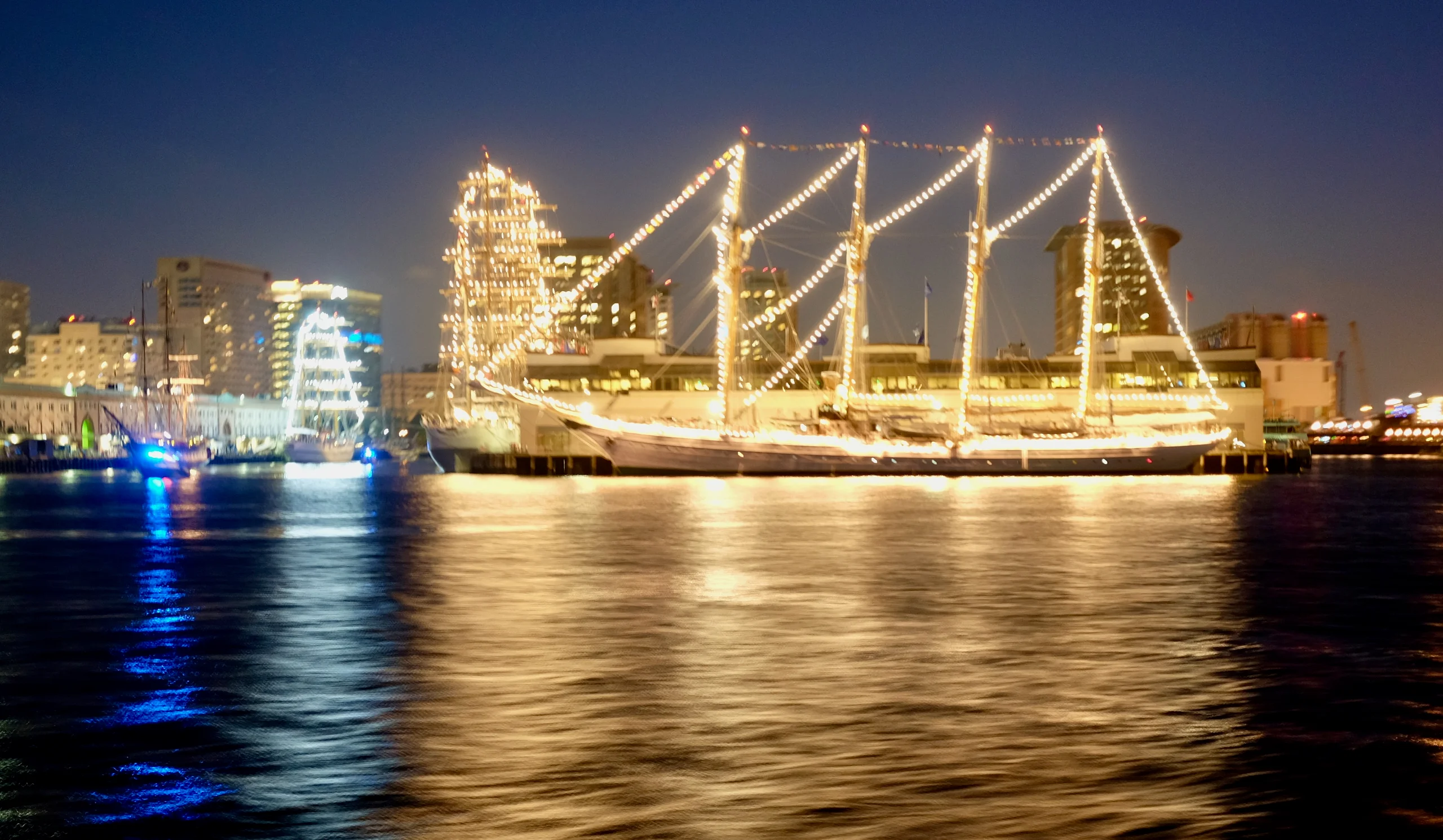 Two tall ships strung with lights sit at a Boston harbor pier at dusk, their reflections streaking across dark water before a lit skyline.
